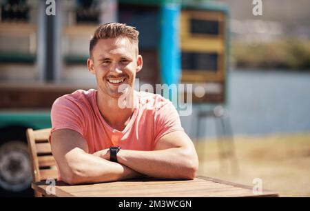 Ich bin heute ganz allein. Porträt eines fröhlichen Mannes, der tagsüber alleine an einem Tisch neben einer Strandpromenade sitzt. Stockfoto