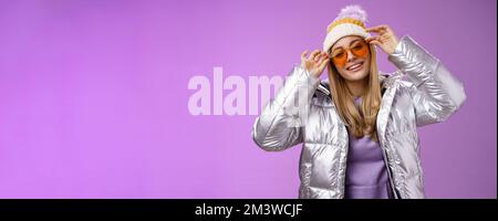 Stilvolle freche gewagte blonde europäische Mädchen handeln cool tragen stilvoll Sonnenbrille Silber Jacke Winter Hut Überprüfung Rahmen Nase kippbaren Kopf Frech lächelnd Stockfoto