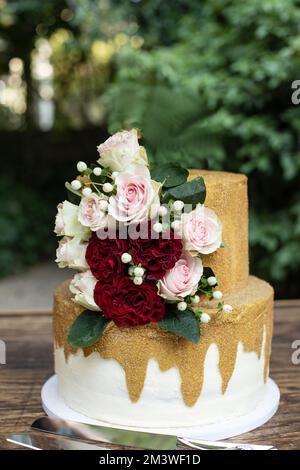 Zweistufige Hochzeitstorte in Weiß und Gold mit roten und pinkfarbenen Rosen und Zruskusblättern Stockfoto