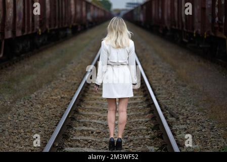 Rückansicht einer attraktiven blonden Frau mittleren Alters in einem weißen Trenchcoat, die auf Gleisen vor Eisenbahnwaggons steht Stockfoto