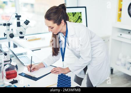 Machen Sie sich Notizen. Eine fokussierte junge Wissenschaftlerin macht Notizen in einem Buch, während sie im Laufe des Tages Tests in einem Labor durchführt. Stockfoto