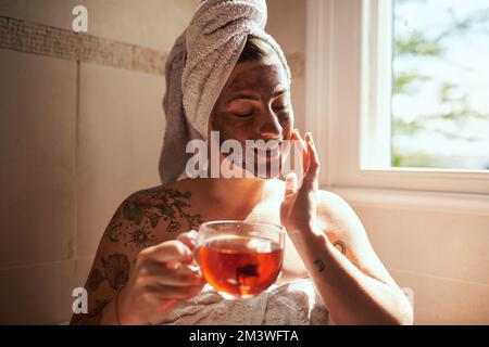 Manchmal muss man sich selbst an erste Stelle setzen. Eine junge Frau, die Tee trinkt, während sie sich zu Hause eine Gesichtsbehandlung gibt. Stockfoto