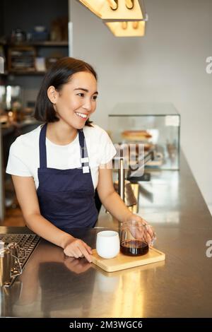 Vertikale Aufnahme eines lächelnden asiatischen Baristas, eines Mädchens, das im Café arbeitet, dem Kunden Befehle erteilt, eine Portion Kaffee zubereitet, Filterkaffee Stockfoto