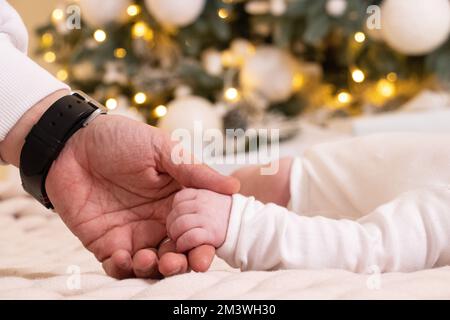 Dads Hand hält die Hand eines Neugeborenen unter dem Neujahrsbaum, die Liebe, die Hand eines Neugeborenen Stockfoto