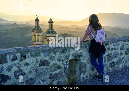 Frau, die den Sonnenuntergang über den Bergen von Grazalema und den Türmen der Kirche des Dorfes Olvera, Cadiz, beobachtet. Stockfoto