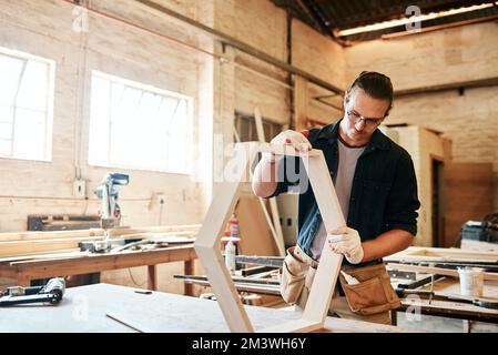 Er hat die Dinge fest im Griff. Ein hübscher junger Zimmermann, der in einer Werkstatt an einem Holzrahmen arbeitet. Stockfoto