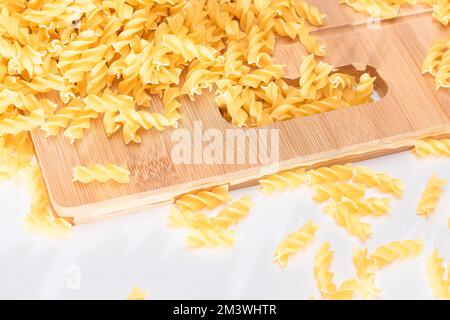 Ungekochte Fusilli Pasta auf Holzbrett und weißem Tisch verstreut. Rohe und trockene Macaroni. Fett und ungesunde Lebensmittel Stockfoto