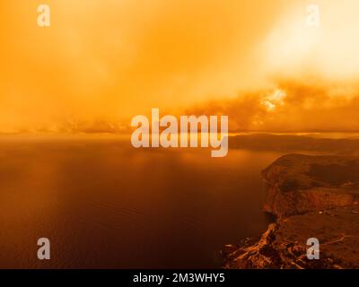 Blick aus der Vogelperspektive auf den roten Sonnenuntergang über dem Meer mit felsigen vulkanischen Klippen. Abstrakte Natur Sommer Sonnenuntergang Meereshintergrund. Kleine Wellen auf goldenem Warm Stockfoto