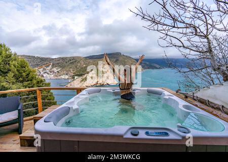 Whirlpool im Freien mit Blick auf die Berge und das Meer. Eine Frau im schwarzen Badeanzug entspannt sich im Hotelpool und bewundert die Aussicht Stockfoto