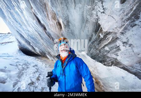 Porträt eines lächelnden alten bärtigen Mannes mit Action-Kamera in blauem Kostüm nahe der Eishöhlenwand am Gletscher im schneebedeckten Bergtal in Almaty, K Stockfoto