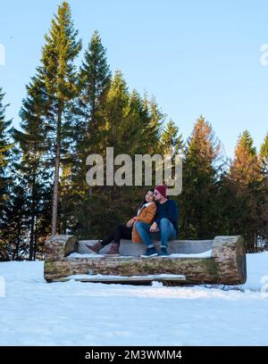 Ein Paar sitzt auf einer Holzbank im Schnee, weiße Männer und asiatische Frauen im Schnee mit Holz im Hintergrund im Harz-Nationalpark in Deutschland. Stockfoto