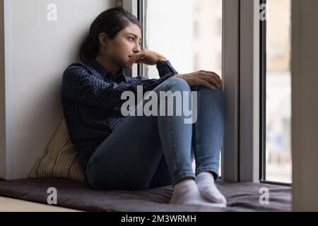 Deprimierte, aufgewühlte Indianerin, die auf einer weichen Fensterbank ruht Stockfoto