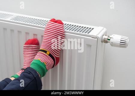 Kinderfüße in warmen Socken auf einem Heizkörper. Ein Mädchen, das sich in einer kalten Wohnung aufwärmt Stockfoto