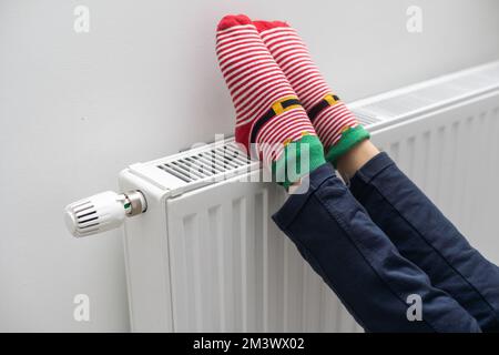 Füße der Person in hellen Wintersocken in der Nähe des Heizkörpers zu Hause Stockfoto