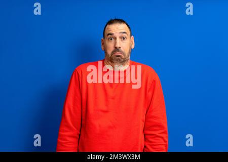 Frontales Porträt eines bärtigen Latino-Mannes in einem roten Pullover, der bezweifelt, isoliert auf blauem Studiohintergrund zu zucken Stockfoto