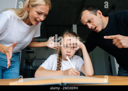 Porträt eines müden kleinen Mädchens, das Hausaufgaben macht, zu Hause am Schreibtisch sitzt, im Hintergrund wütender Eltern, die zusammen faule Tochter schimpfen. Stockfoto