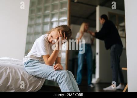 Porträt eines traurigen, einsamen, weinenden kleinen Mädchens, das auf dem Sofa sitzt, während Eltern sich im Wohnzimmer im Hintergrund streiten und streiten. Stockfoto