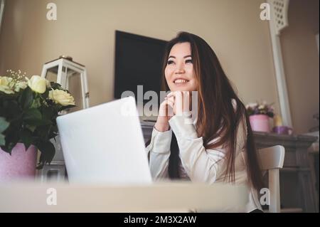 Junge, wunderschöne asiatische Geschäftsfrau, die online arbeitet, mit einem Laptop, der am Tisch im Heimbüro aufgestellt wurde. Eine lächelnde Freiberuflerin hat Fernarbeiten im Wohnzimmer mit weißen Rosenblumen Stockfoto