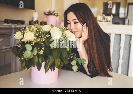 Eine wunderschöne asiatische Frau sitzt am Tisch im Wohnzimmer. Die helle Inneneinrichtung bewundert den Blumenstrauß der weißen Rosen. Lächelnde junge Dame, die ein Überraschungsgeschenk erhalten hat Stockfoto