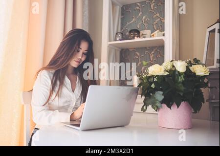 Junge, wunderschöne asiatische Geschäftsfrau, die online arbeitet, mit einem Laptop, der am Tisch im Heimbüro aufgestellt wurde. Eine lächelnde Freiberuflerin hat Fernarbeiten im Wohnzimmer mit weißen Rosenblumen Stockfoto