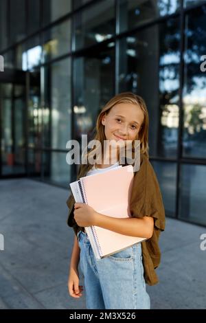 Lächelndes Mädchen, das auf der Straße steht und Notizbücher hält. Kinder im Freien. Zurück zum Schulkonzept Stockfoto