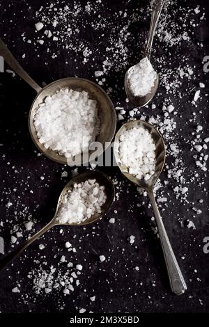 Draufsicht von vier Löffeln mit grob körnigem Meersalz auf schwarzem Hintergrund. Flach verlegt. Draufsicht. Speisekonzept. Dunkle Stimmungsfotografie. Stockfoto