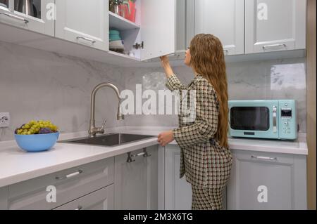 Süße junge Hausfrau öffnet die Tür zum Küchenschrank Stockfoto