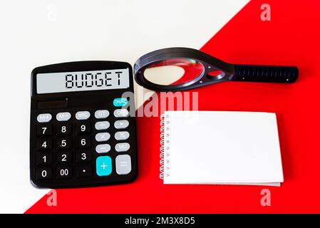 Rechner mit dem Wort BUDGET auf dem Display. Vergrößerungsglas und Notizblock auf rot-weißem Hintergrund. Konzeptfoto Stockfoto