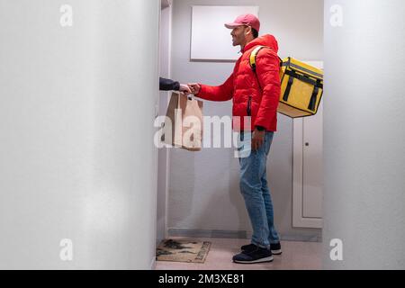 Das Konzept der Lebensmittellieferung. Ein junger, lächelnder Afroamerikaner mit einem Rucksack, der Produkte vor der Tür liefert. Stockfoto