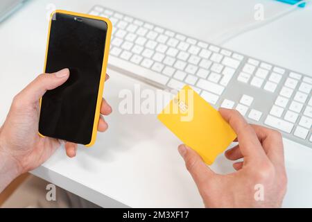 Ein unbekannter Mann, der online mit Kreditkarte und Handy vor einem Computer bezahlt. Stockfoto