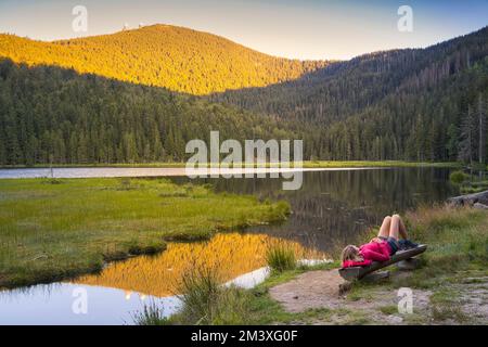 Eine europäische Frau liegt auf einer Holzbank am wunderschönen kleinen Arbersee bei Sonnenuntergang. Blick auf den Großer Arber mit seinen zwei Türmen. Bayerischer Wald Stockfoto