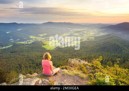 Goldene Stunde. Eine weiße Frau, die auf dem Osser-Berg sitzt und die Landschaft von Lamer Winkel mit der Kleinstadt Lam überblickt. Berg Hohenbogen rechts Stockfoto