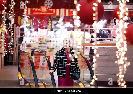 Eine schöne, reife Frau steht auf der Rolltreppe im Einkaufszentrum vor dem Hintergrund von Neujahrs- oder Weihnachtsdekor Stockfoto