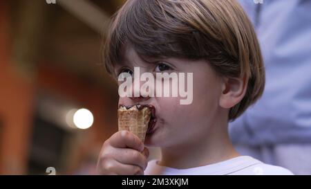Ein kleiner Junge genießt draußen auf der Straße Eiscreme. Porträtgesicht eines Kindes, das Konus isst Stockfoto