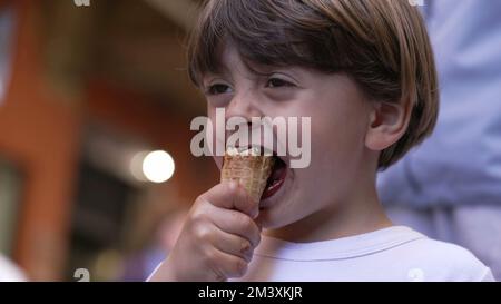 Ein kleiner Junge genießt draußen auf der Straße Eiscreme. Porträtgesicht eines Kindes, das Konus isst Stockfoto