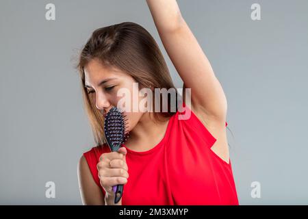 Junge Frau in rotem Kleid singt leidenschaftlich und tut so, als wäre ihre Haarbürste ein Mikrofon, sie ist sehr in der Rolle und glücklich Stockfoto