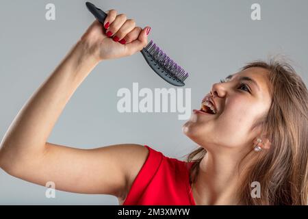 Junge Frau in rotem Kleid singt leidenschaftlich und tut so, als wäre ihre Haarbürste ein Mikrofon, sie ist sehr in der Rolle und glücklich Stockfoto