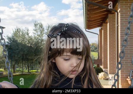 Bild eines bezaubernden kleinen brünetten Mädchens mit Haaren, die im Wind wehen und zu Hause auf der Schaukel im Garten spielen. Stockfoto