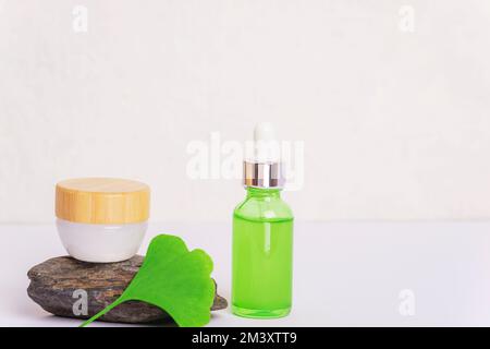 Frisches grünes Ginkgo-Biloba-Blatt, kosmetische Serumflasche und Cremebecher auf weißem Hintergrund. Großaufnahme, Kopierraum. Stockfoto