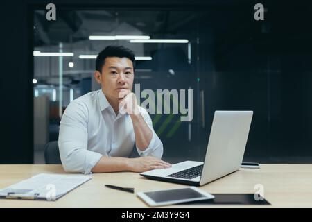 Porträt eines jungen, gut aussehenden, erfolgreichen Asiaten, Geschäftsmann, Firmengründer, Büroangestellten, der an einem Schreibtisch im Büro sitzt, selbstbewusst in die Kamera schaut und seinen Kopf mit der Hand hält. Stockfoto