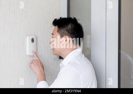 Nahaufnahme. Ein junger Asiate ruft mit dem Finger an der elektronischen Türklingel an der Wand des Hauses an, Büro. Spricht in die Sprechanlage Stockfoto