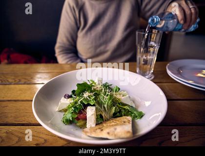 Grüner Salat mit Fetakäse auf einer weißen Keramikplatte und eine Frau gießt Wasser in ein Glas auf dem Hintergrund Stockfoto