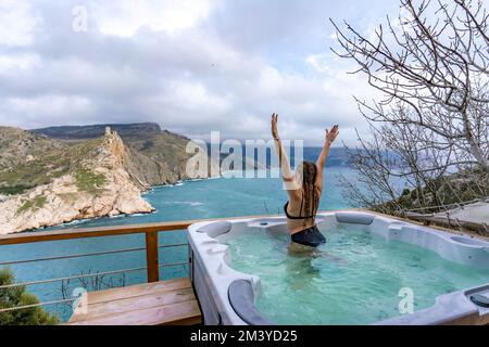 Whirlpool im Freien mit Blick auf die Berge und das Meer. Eine Frau im schwarzen Badeanzug entspannt sich im Hotelpool und bewundert die Aussicht Stockfoto