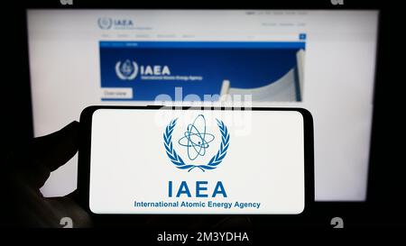 Person, die ein Smartphone mit dem Logo der Internationalen Atomenergie-Organisation (IAEO) auf dem Bildschirm vor der Website hält. Konzentrieren Sie sich auf das Display des Telefons. Stockfoto