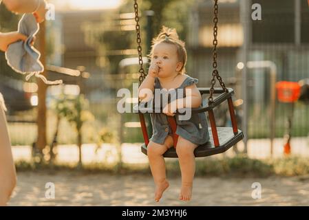 Ein Kleinkind in einem Kleid saugt an einem warmen Sommerabend den Daumen an einer Schaukel Stockfoto