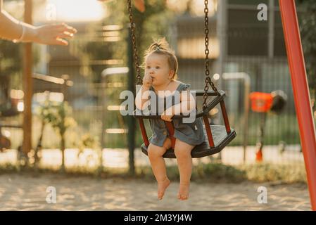 Ein Kleinkind in einem Kleid saugt an einem warmen Sommerabend den Daumen an einer Schaukel Stockfoto