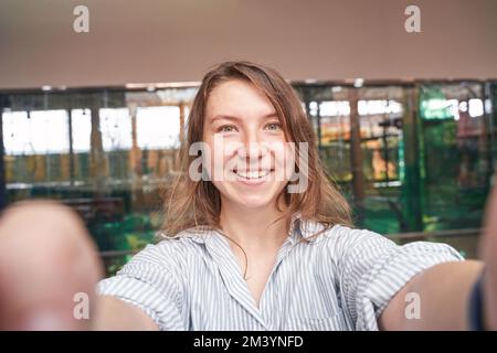 Young woman doing self portrait indoors. Blogger selfie video. Happy emotion. Smiling female person. Lifestyle action. Pretty posing. Copyspace. Holding phone at two hands Stock Photo