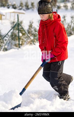 Ein Mädchen schaufelt Schnee auf der Heimfahrt. Schöner verschneiter Garten oder Vorgarten. Ein Teenager, der im Winter mit einer Schaufel Schnee entfernt Stockfoto