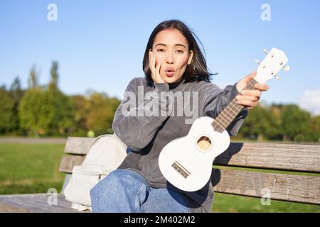 Portrait of young girl musician, sitting in park with ukulele guitar, looking surprised at camera, saying wow