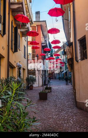 luino, Italien - 12-06-2022: Wunderschöne Straße im historischen Zentrum mit farbenfrohen Geschäften und hängenden roten Sonnenschirmen in Luino Stockfoto